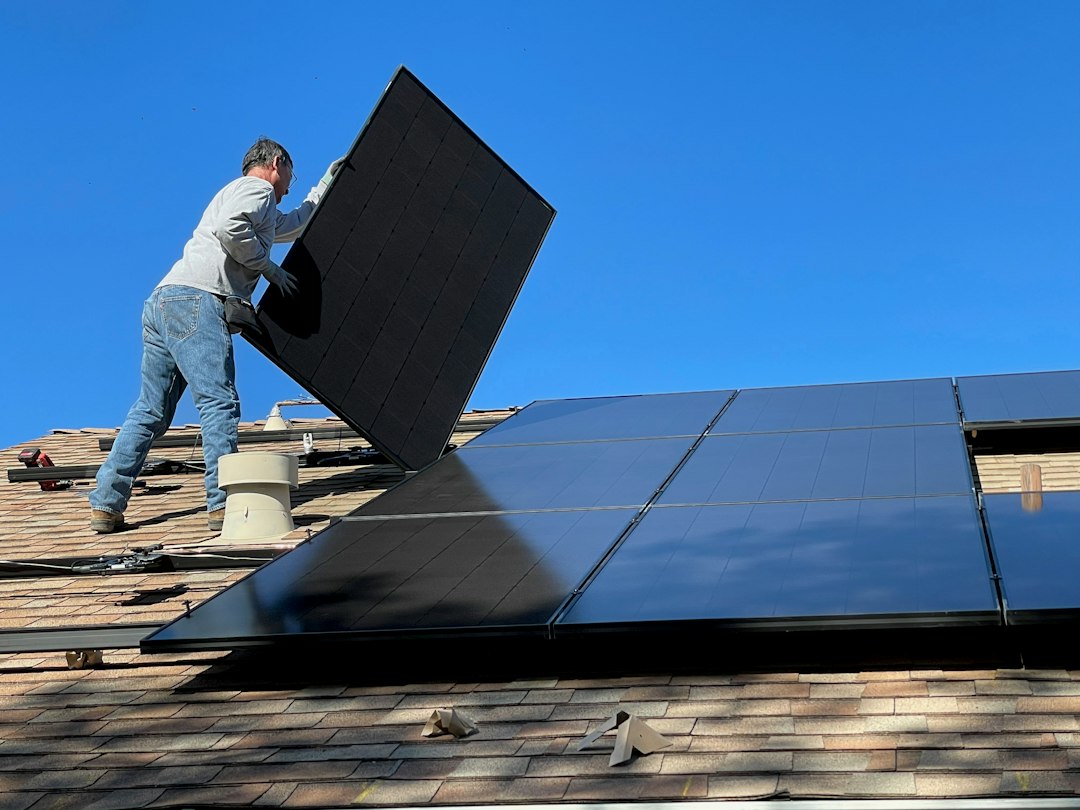 Batería Virtual: Qué Es, Cómo Funciona y con Qué Comercializadoras Contratar: A person installing a black solar panel on a shingled roof under blue sky