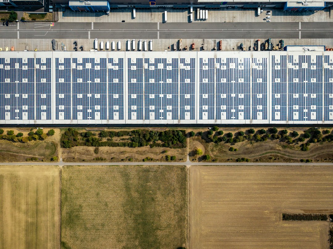 Efecto del Polvo, Polen y Excrementos de Aves en las Pérdidas de Generación: An aerial view of a large warehouse roof covered in solar panels beside fields